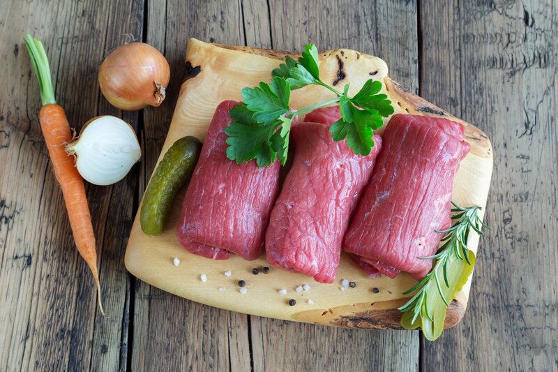 Drei rohe Rinderrouladen auf einem Holzbrett, umgeben von Gewürzgurke, Zwiebel, Möhre und frischen Kräutern – Zutaten für ein klassisches Rouladenrezept | © AdobeStock_196861401_(c)_juefraphoto