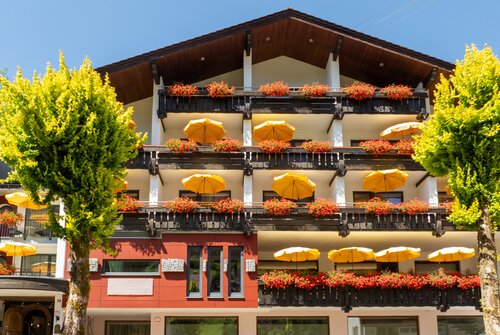 Ringhotel Sonnenhof in Baiersbronn, ein 4-Sterne-Hotel im Schwarzwald, mit Balkonen mit leuchtend gelben Sonnenschirmen und roten Blumen an einer traditionellen Holzfassade. | © Ringhotels Sonnenhof in Baiersbronn